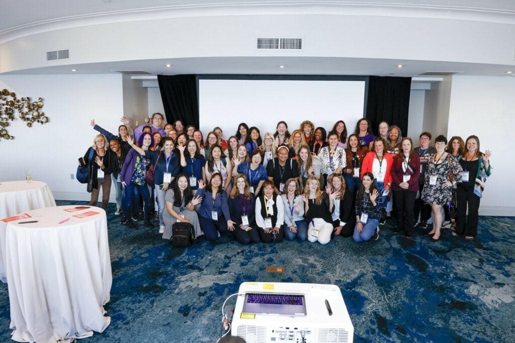 Large group of women gathered in front of a projector, smiling for the camera at NANOG 89's Women in Tech event.